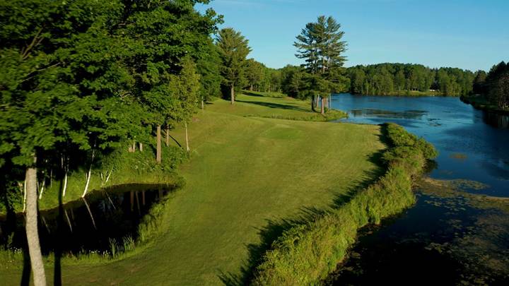 Lush green golf course bordered by a serene lake and tall trees under a clear blue sky, inviting outdoor leisure and recreation.
